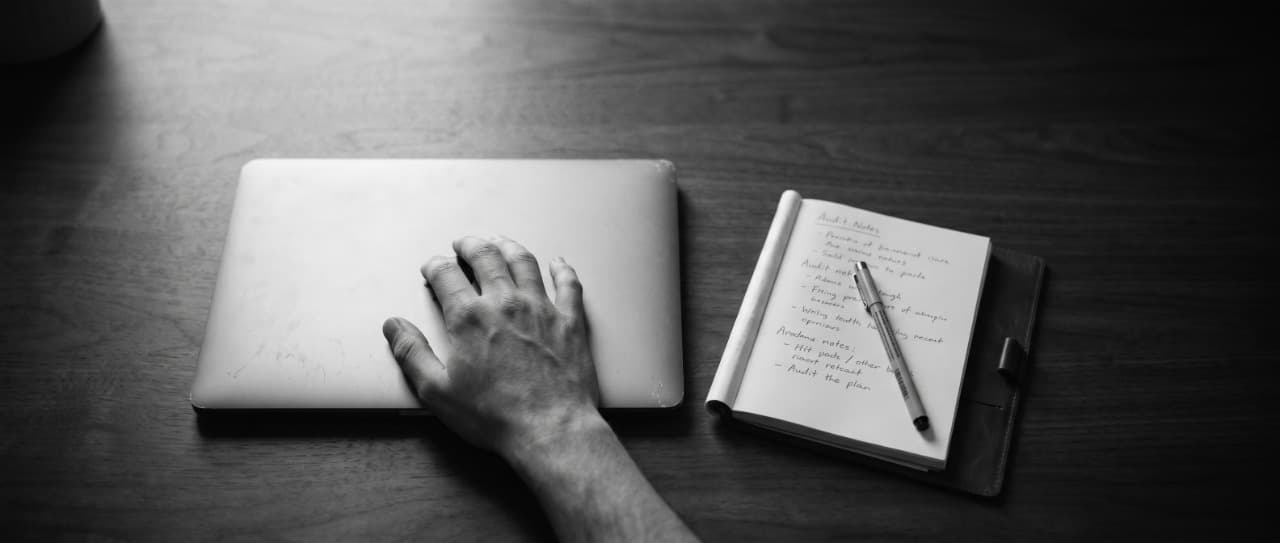 A hand resting on a closed laptop next to an open notebook of handwritten audit notes on a dark wooden desk